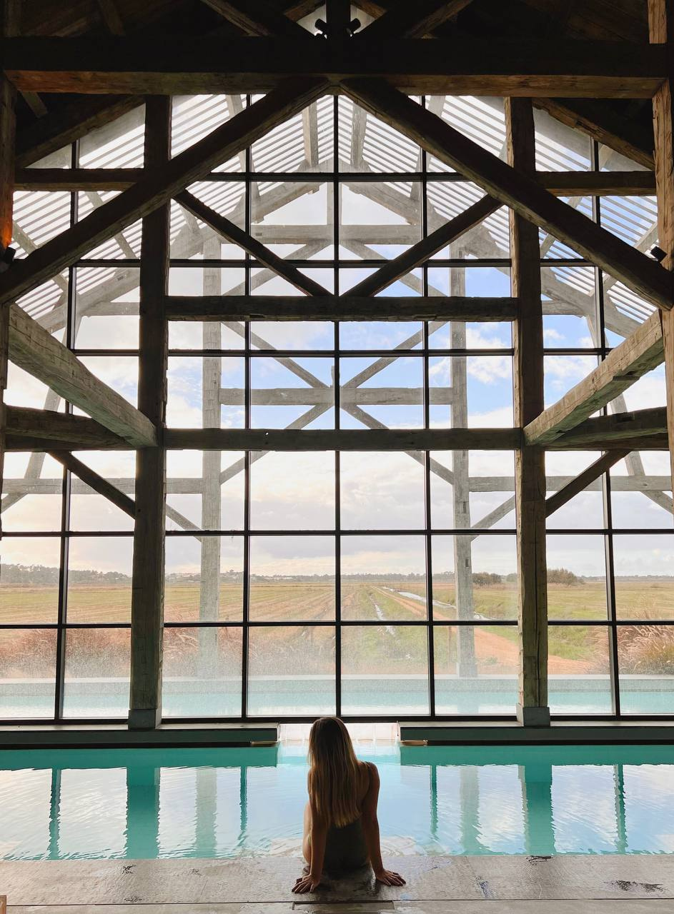 Woman sitting at the edge of an indoor pool inside a timber-framed barn overlooking rice fields