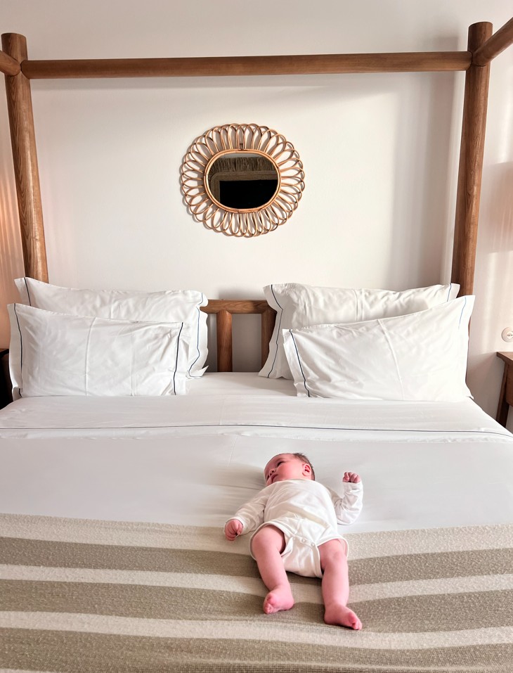 Newborn baby lying on a white hotel bed with a wooden four-poster frame in Comporta