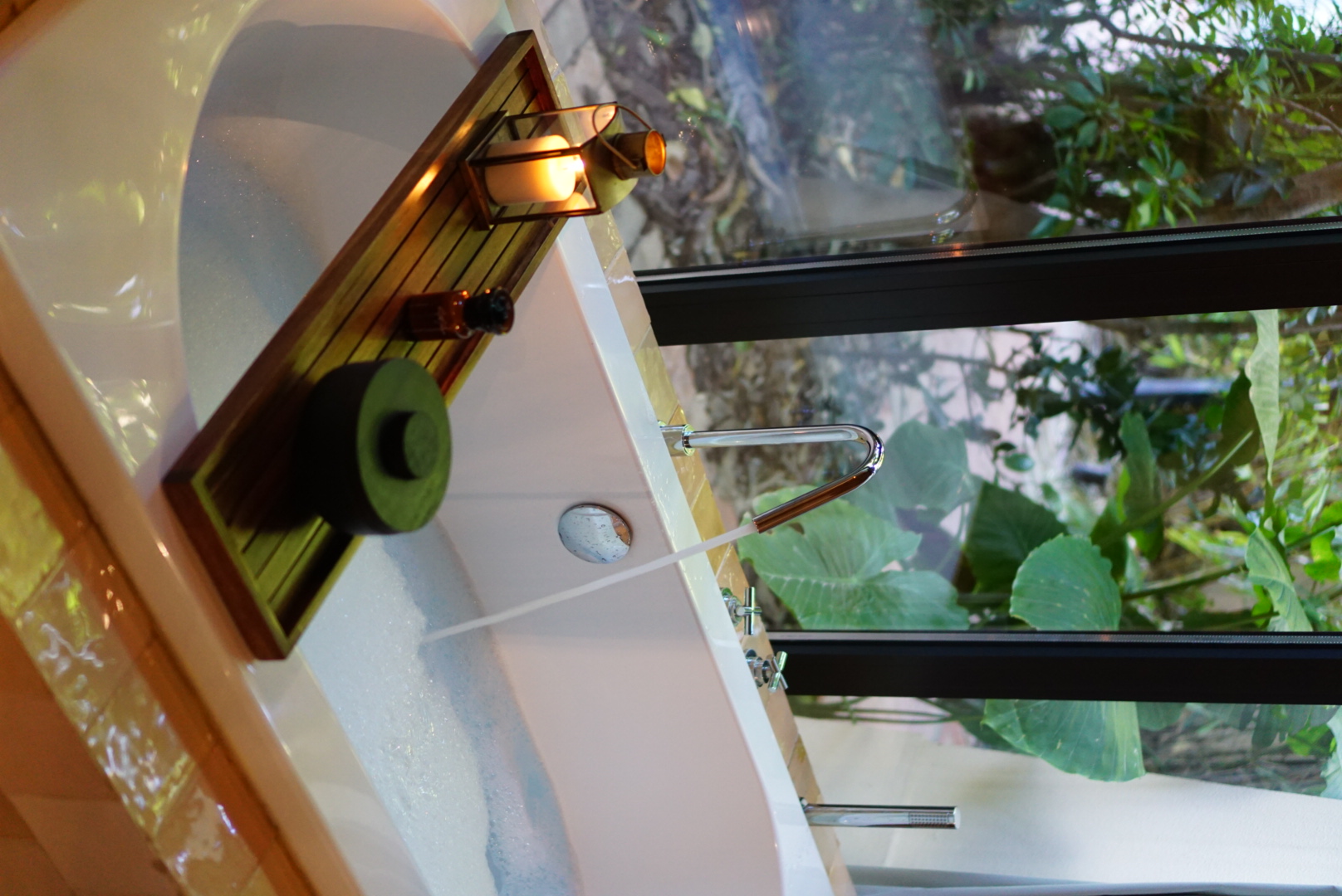 Bathtub filling with water beside a window overlooking tropical greenery, with a candle on a wooden tray